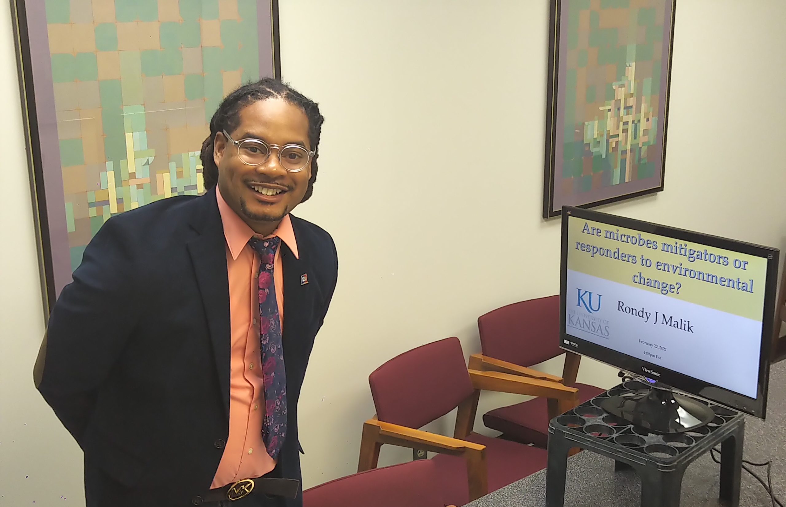 A brown skinned man wearing glasses, a black jacket, a light pink dress shirt, and a colorful tie poses smiling next to a device displaying a presentation slide