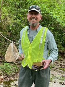 Certified Senior Ecologist Kris Hallinger poses in the field, holding a net and a rock amidst natural surroundings