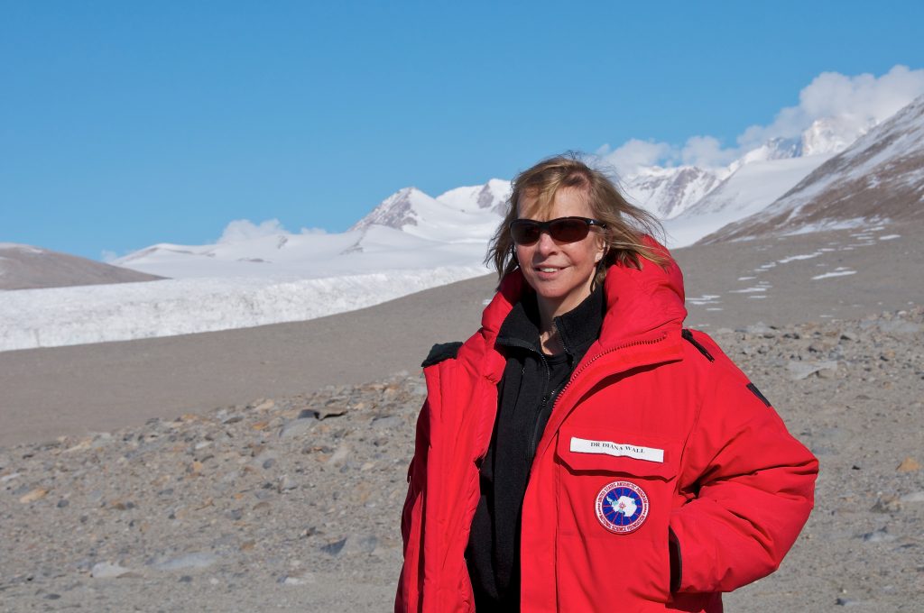 Diana Wall wears a red parka and sunglasses against the backdrop of an Antarctic landscape.