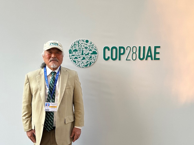 Dennis Ojima poses next to the COP28 Logo on a wall.