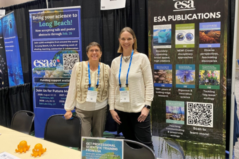 Two women, Catherine O'Riordan and Adrienne Sponsberg, pose at the ESA booth at the Ocean Sciences Meeting; they are flanked by promotional banners, one reading "Bring your science to Long Beach" and the other "ESA publications"