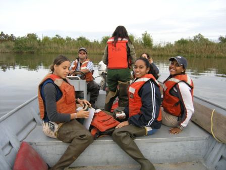 A group of young ecologists wear life jackets sitting in an aluminum boat on calm water with reeds in the background.