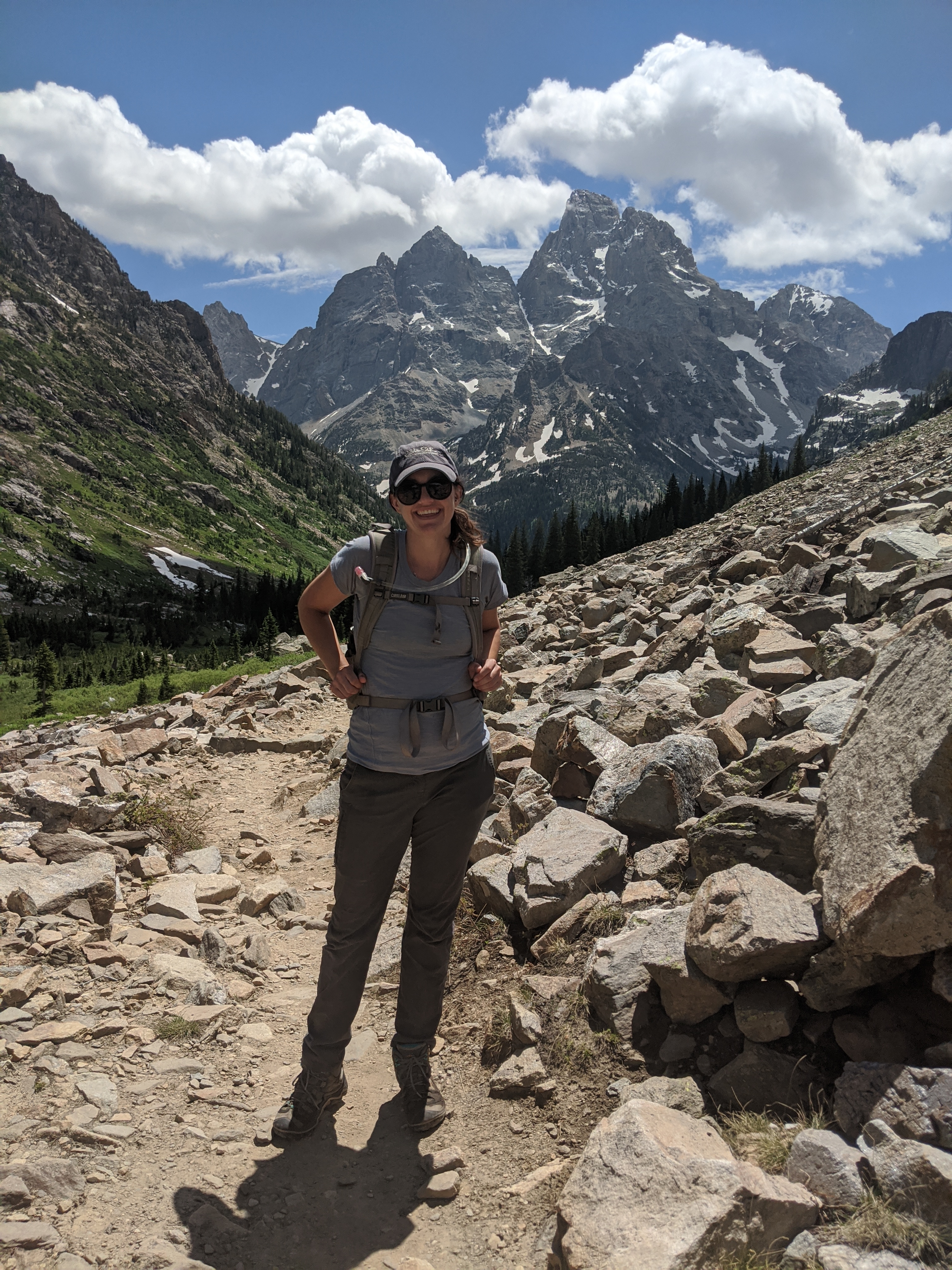 A 2021 Scientists in Park Fellow smiles for the camera with hiking gear on in front of her site in a rocky mountain chain.