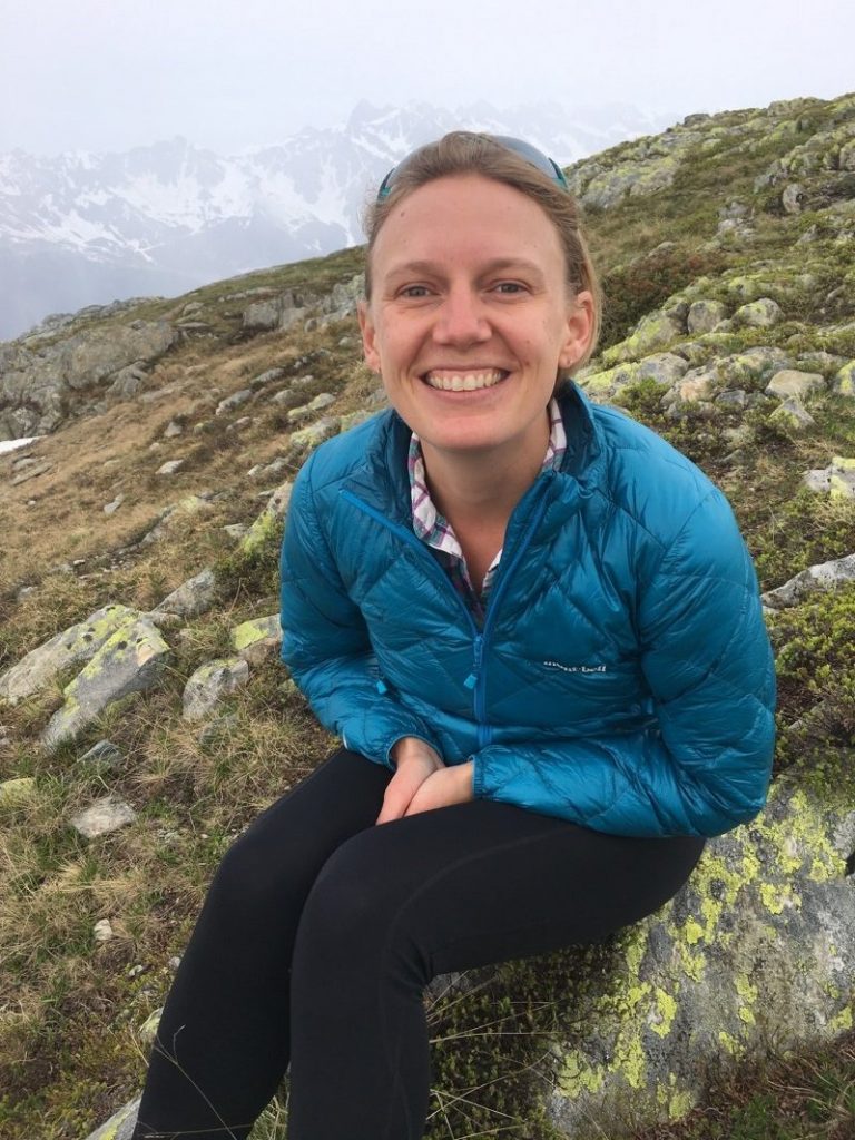 Smiling woman sitting on lichen-covered rocks; clearly in high-alpine environment (snow-covered mountain peaks visible in background)