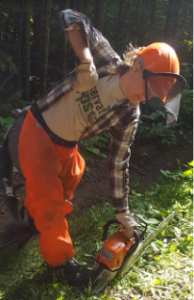 A woman standing over a chainsaw that she is starting. She is wearing orange pants and an orange hard hat with a visor, and a plaid shirt over a tan t-shirt.