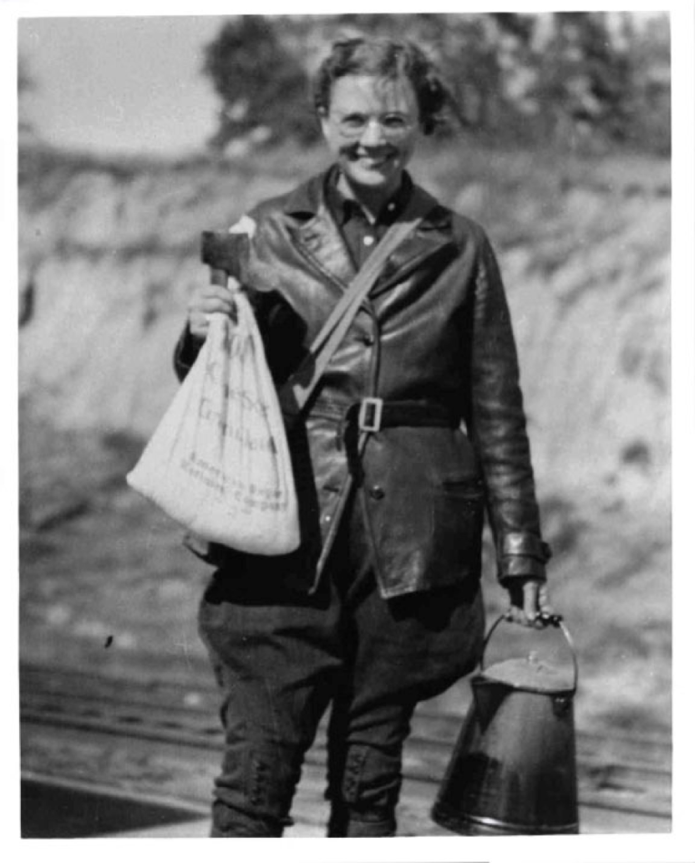 Dr. Mary Talbot stands with an axe and burlap bag in her right hand and what appears a water jug to be a metal container hanging from a handle in her left hand. Black and white.