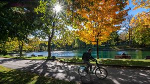 A gentleman rides along a path in a park with changing foliage and a lake nearby.