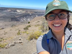 The author of this post, Sana, smiling with a green Scientists in Park hat. The Kilauea caldera is seen behind her.
