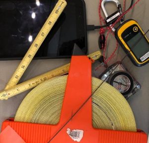 a tablet, compass, GPS unit, large field measuring tape, and folding ruler lay together in a loose pile