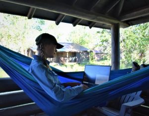  woman with braids sits in a blue hammock working on a computer