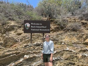 Intern standing in front of sign for the Buck Island Reef National Monument with a rocky hillside and desert plants behind her