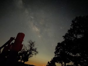 The Milky Way and Stars above the LBJ Ranch. The silhouette of a pole and trees can be seen near the bottom of the image, and an orange glow is present on the horizon.