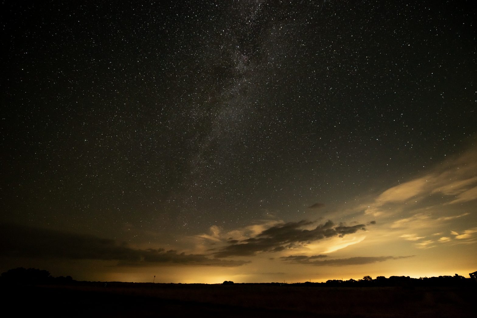 The stars and the Milky Way over the LBJ Ranch. There are some clouds and an orange glow on the horizon.