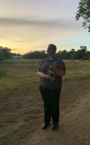 Isabel carrying a water bottle and a clipboard while wearing the Scientists in Parks polo shirt stands on a trail at the Johnson Settlement near sunset.