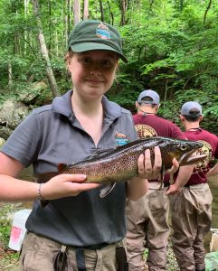 Rowan (author) smiles at the camera as she holds a big, invasive brown trout. The trout in question has an open mouth like it's snarling, and in the background there are two grown men looking at the tree that are set besides the riverbank. 