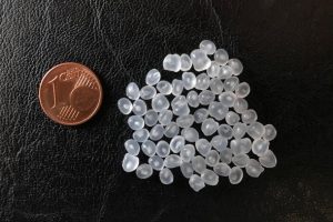 A pile of about 60 small, smooth semi-transparent plastic pellets on a black leather surface. To the left of the pellets is a 1-cent coin for scale. The pile of pellets is about four times larger than the surface of the coin. 