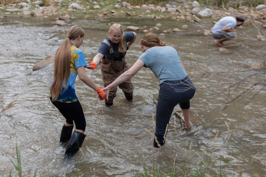 Three students stand holding hands in the rushing creek in waders or field boots. One student is in the background squatting to evaluate something in the creek. 