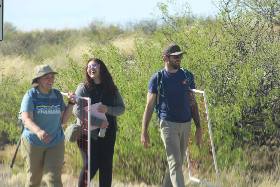 Three students walk next to lush green vegetation smiling and laughing. Two of the students carry sampling quadrats.