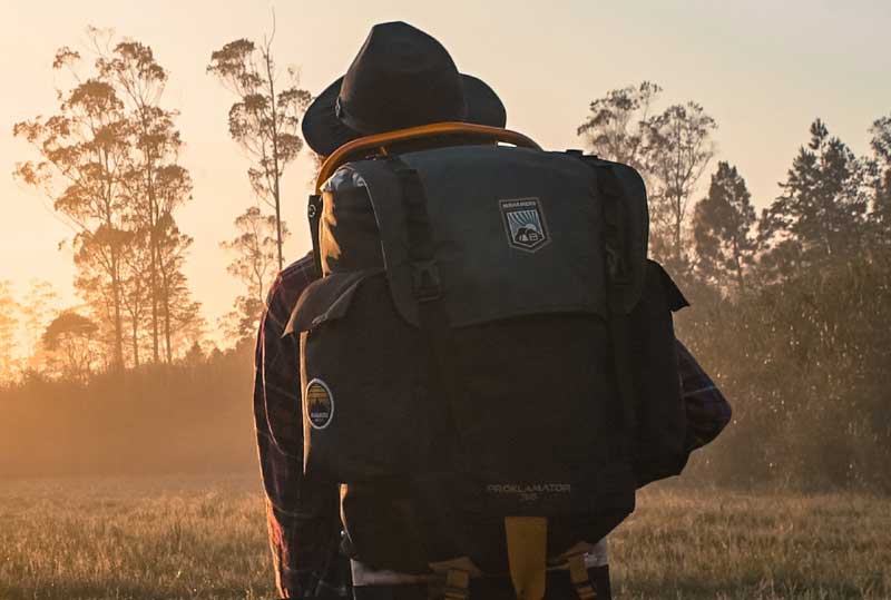 A person stands with their eye fixed to a position on the horizon wearing a full brimed field hat, a full hiking pack and a red flannel shirt.  The setting sun glares at us.